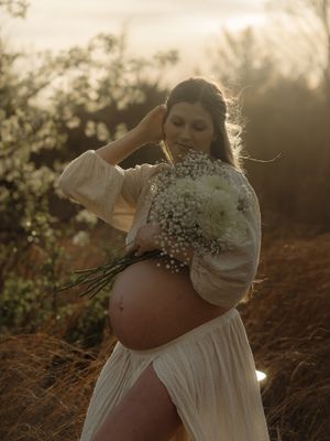 Pregnant woman in white dress holding wildflowers at golden hour in a sunlit outdoor setting.