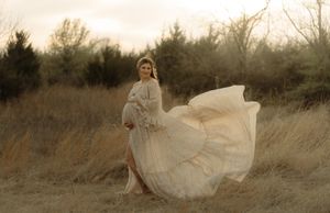 Pregnant woman in flowing ivory boho gown poses outdoors in golden field with wind-swept skirt at sunset.