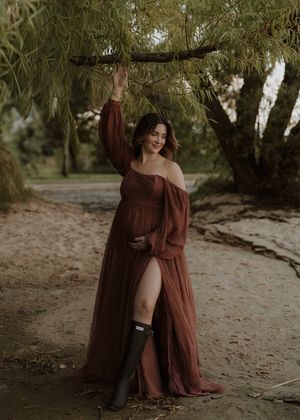 Pregnant woman in rust tulle maternity gown and black boots poses under willow tree branches outdoors.