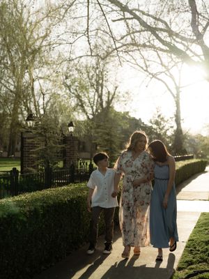 A family of three walks along a sunlit garden path surrounded by lush hedges and trees in golden hour light.