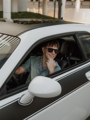 Smiling young man wearing sunglasses sitting in a white sports car with racing stripes, looking out the window.