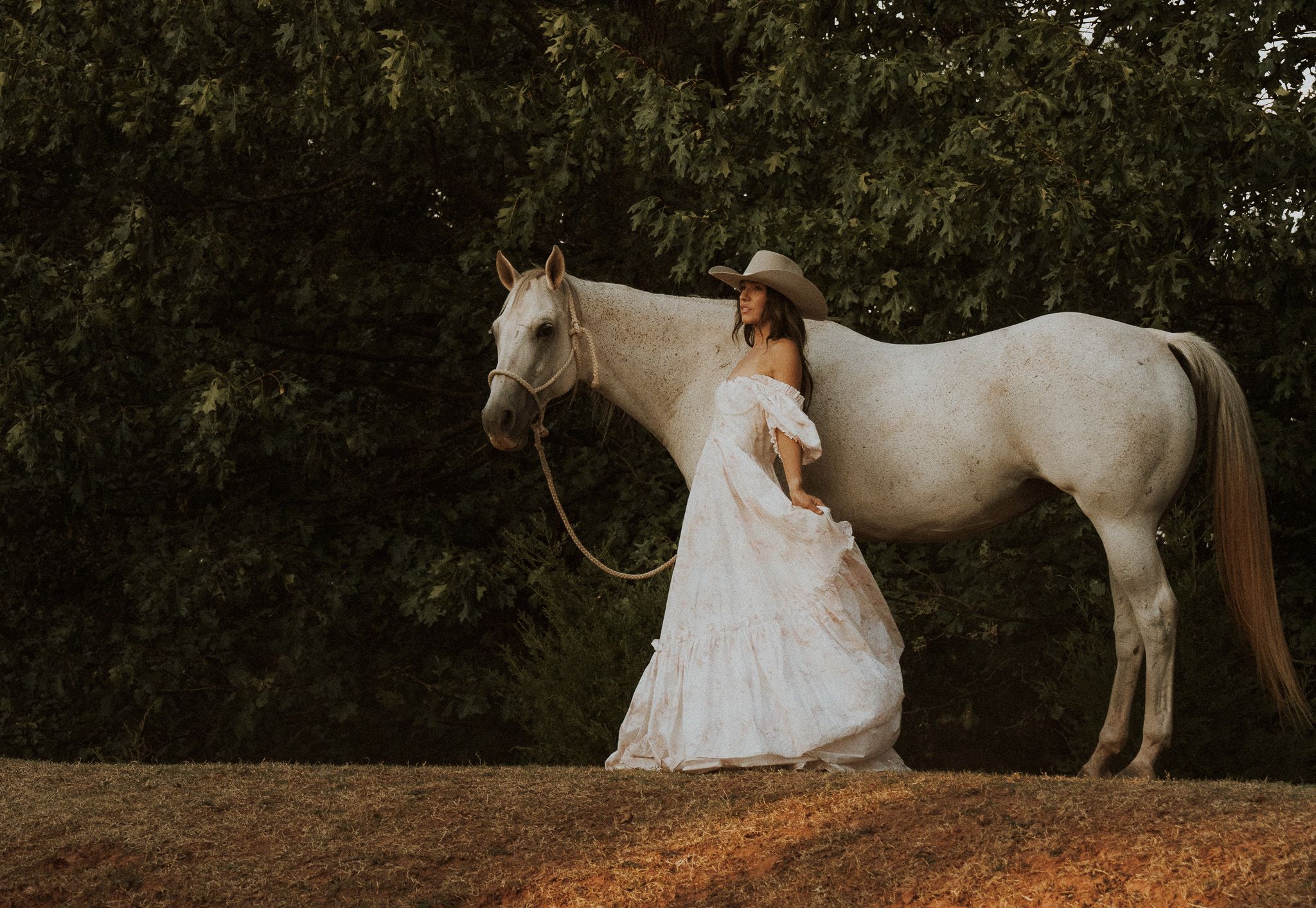 Woman in white dress and cowboy hat stands beside a white horse outdoors at golden hour.