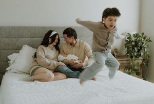 Parents sit on bed with newborn while excited young boy jumps beside them in bright cozy bedroom.