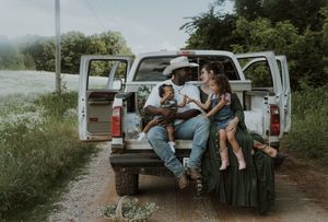 Family sitting on tailgate of white pickup truck on rural dirt road surrounded by green fields and trees.