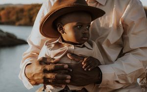 An adult holds a young Black baby wearing a tan hat and beige outfit near a scenic waterway in warm golden light.