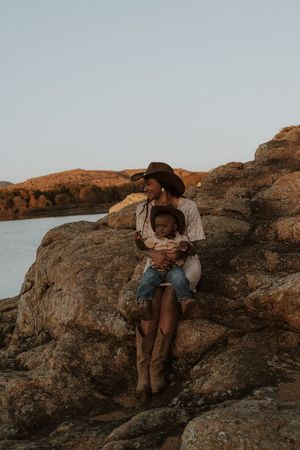 Two people embrace while sitting on rocky coastal cliffs at sunset with ocean views in the background.