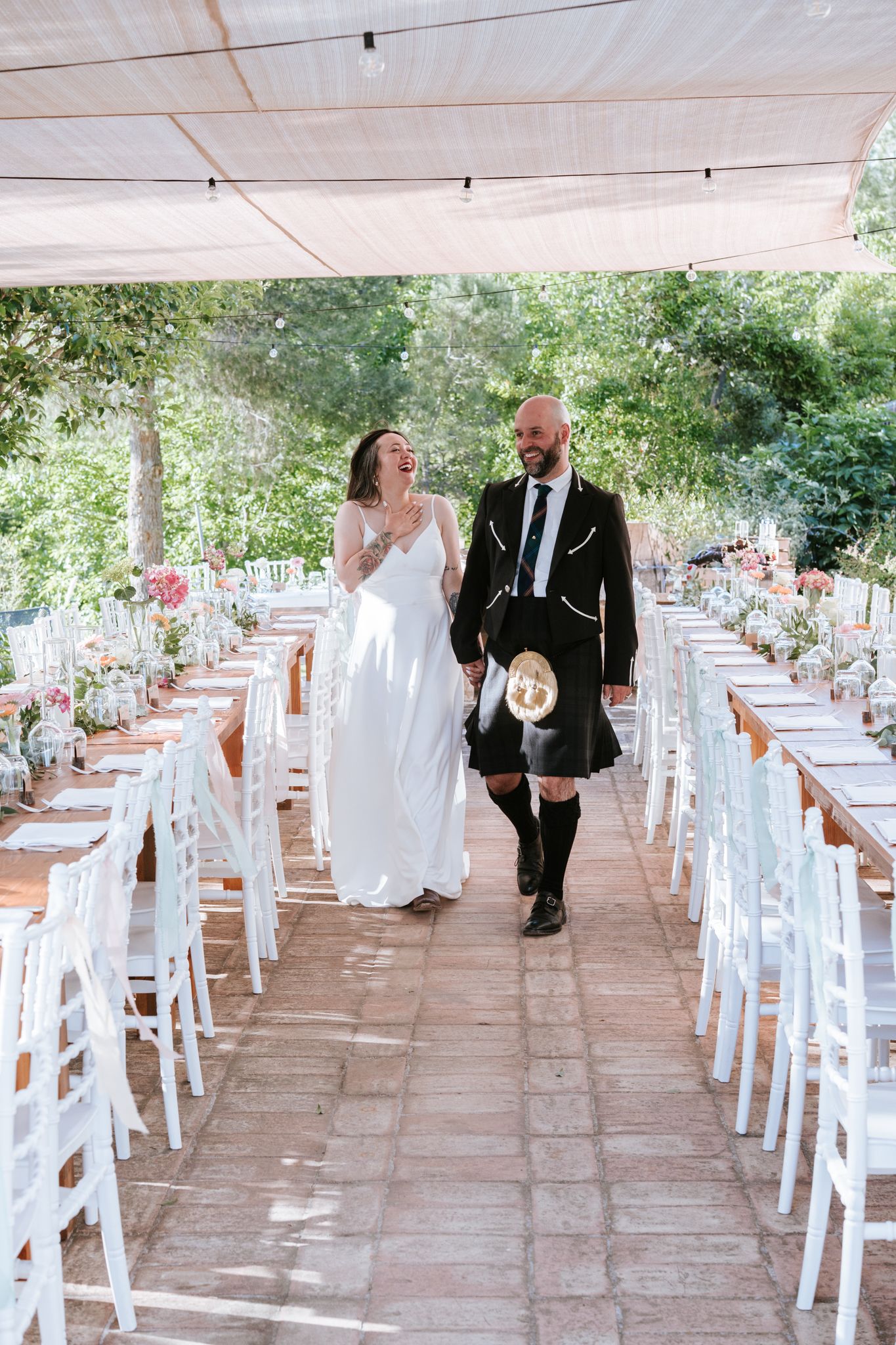 Laughing bride and kilted groom walk through elegantly set outdoor wedding reception dining area.