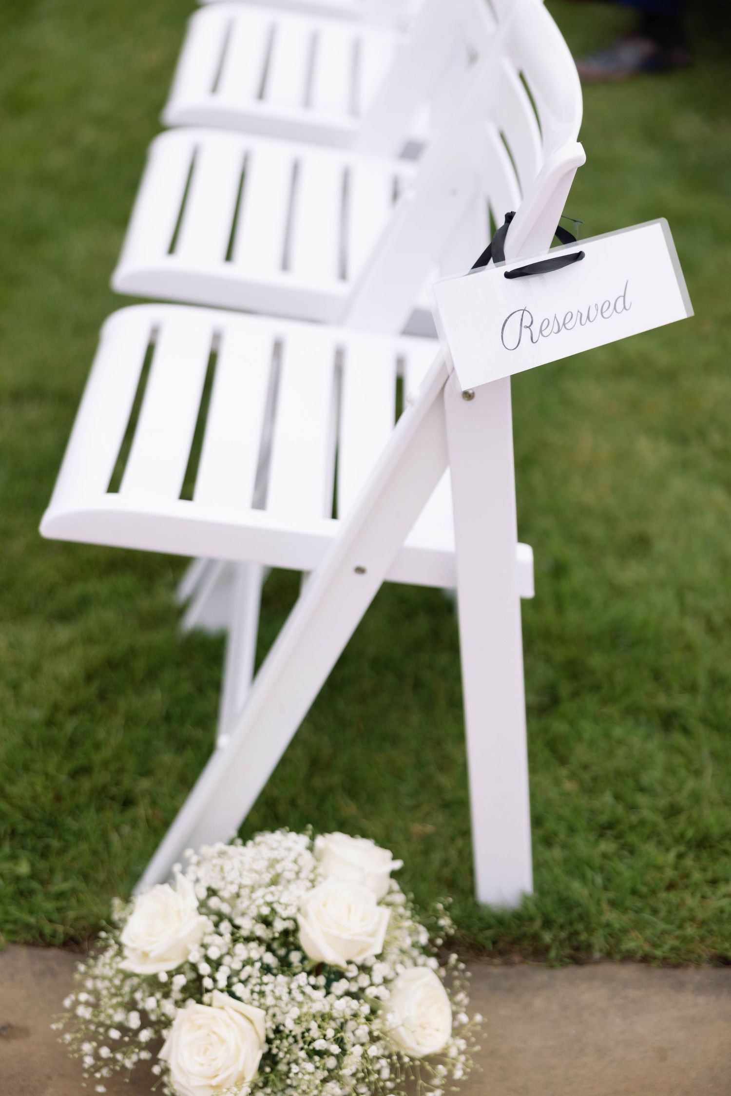White folding chair with reserved sign and white rose bouquet arrangement on grass.