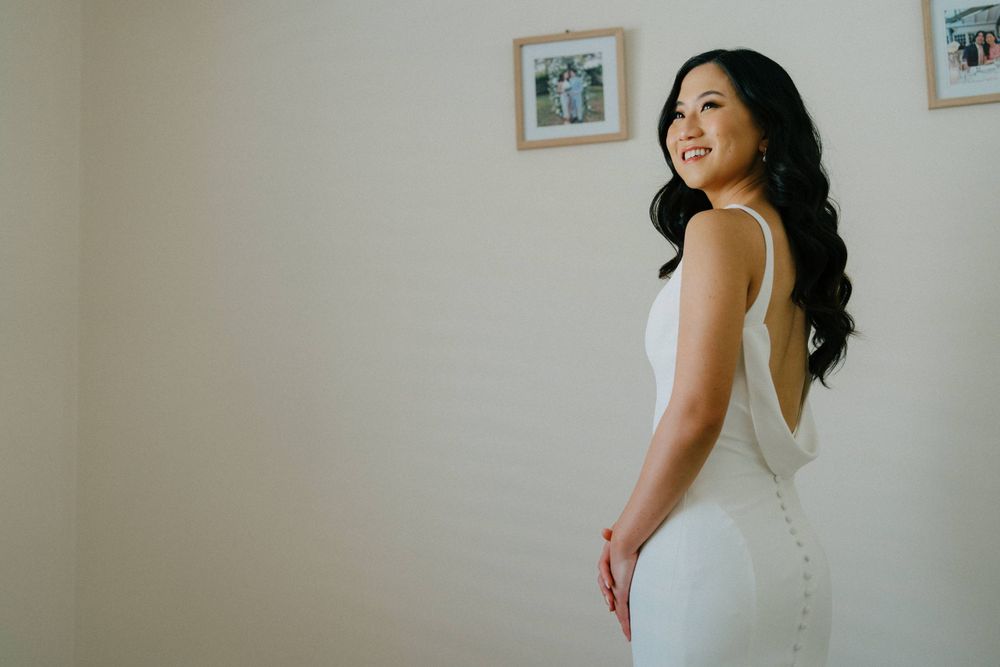 Elegant white wedding dress shown from behind against minimalist wall with framed photos.