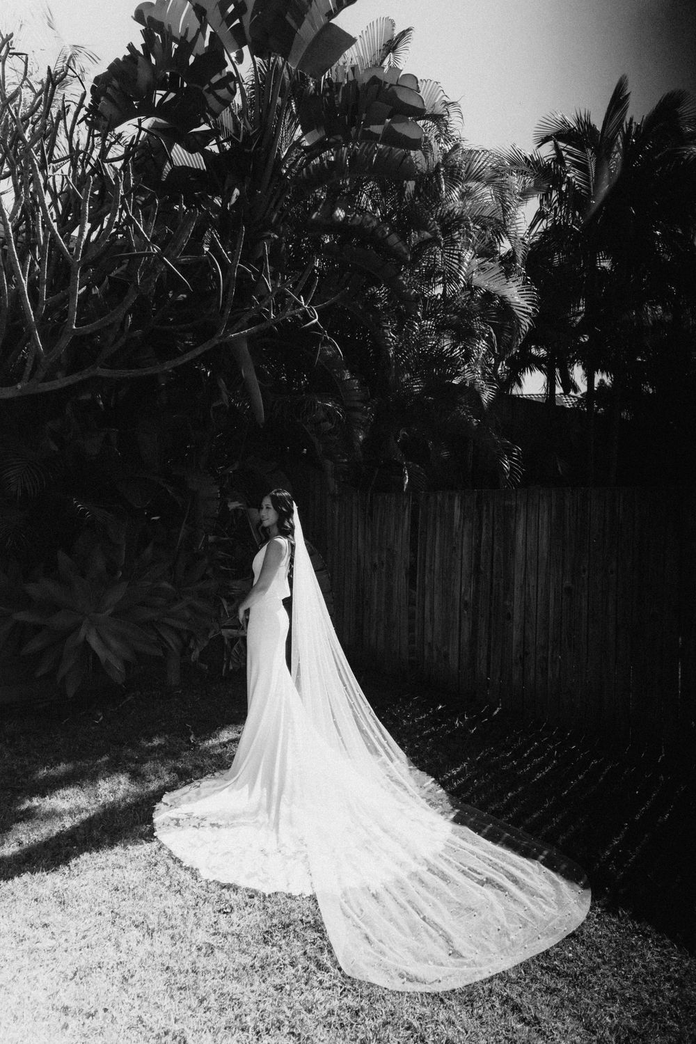 Dramatic black and white photo of a wedding dress with long flowing veil in a garden setting.