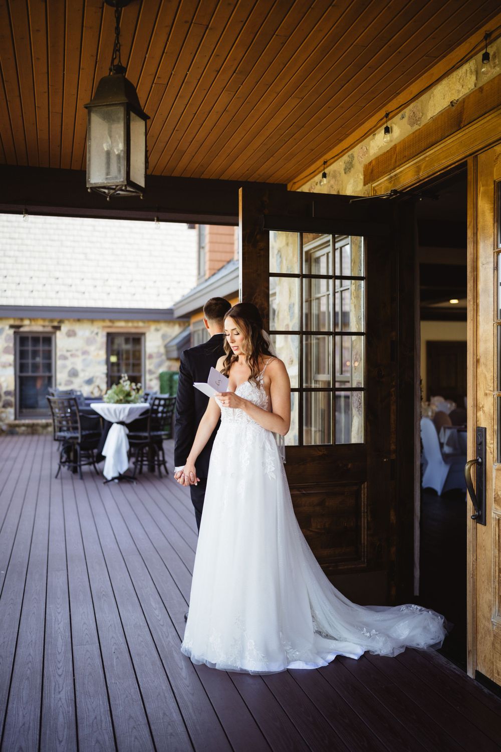 A bride in a flowing white gown reads on a rustic wooden deck with lantern lighting.