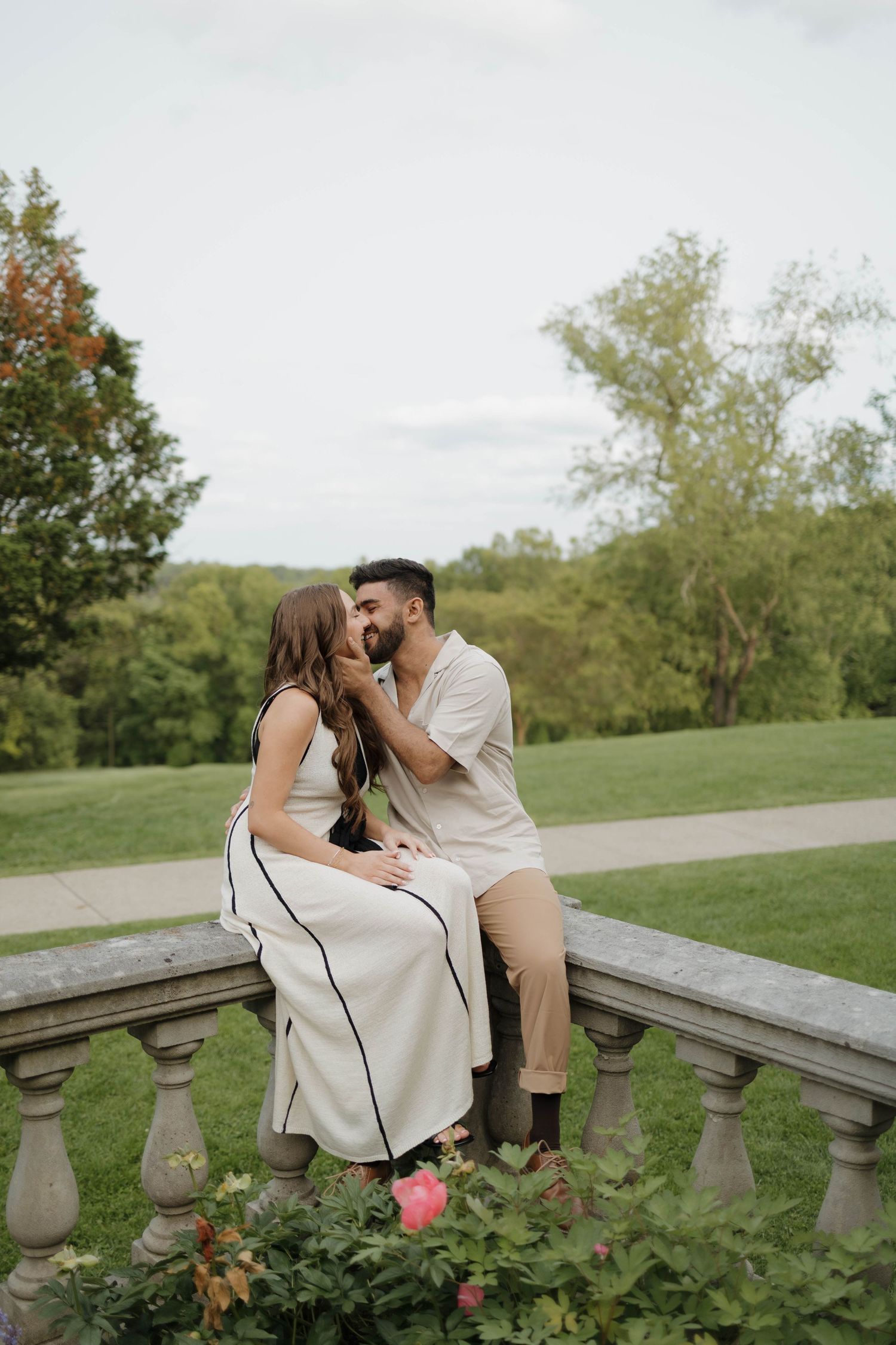 A romantic proposal scene unfolds on a grassy lawn in front of a stone building with someone kneeling and their partner standing.
