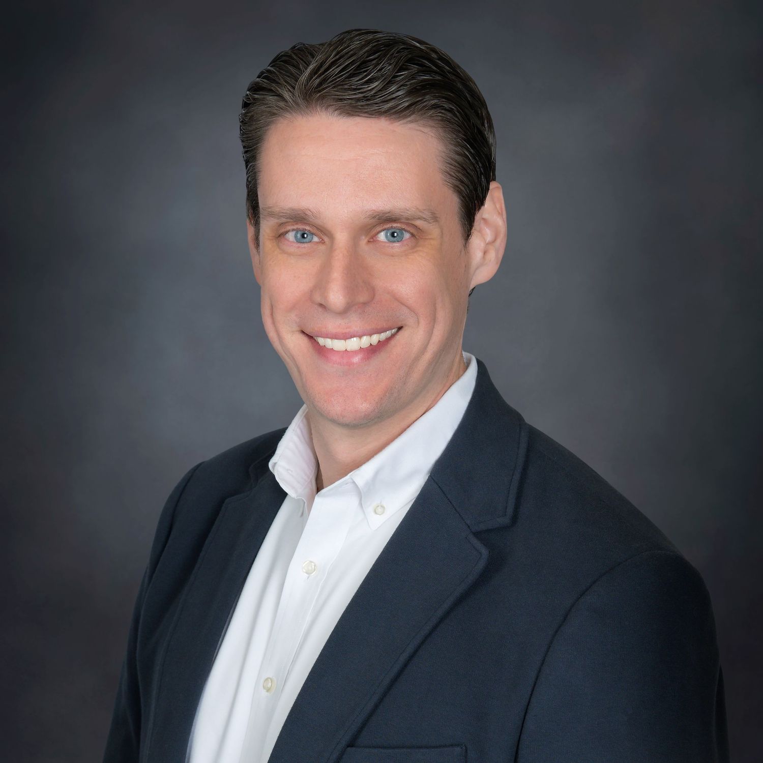 Professional headshot of someone in a grey suit and blue tie with glasses and a beard smiling at the camera.