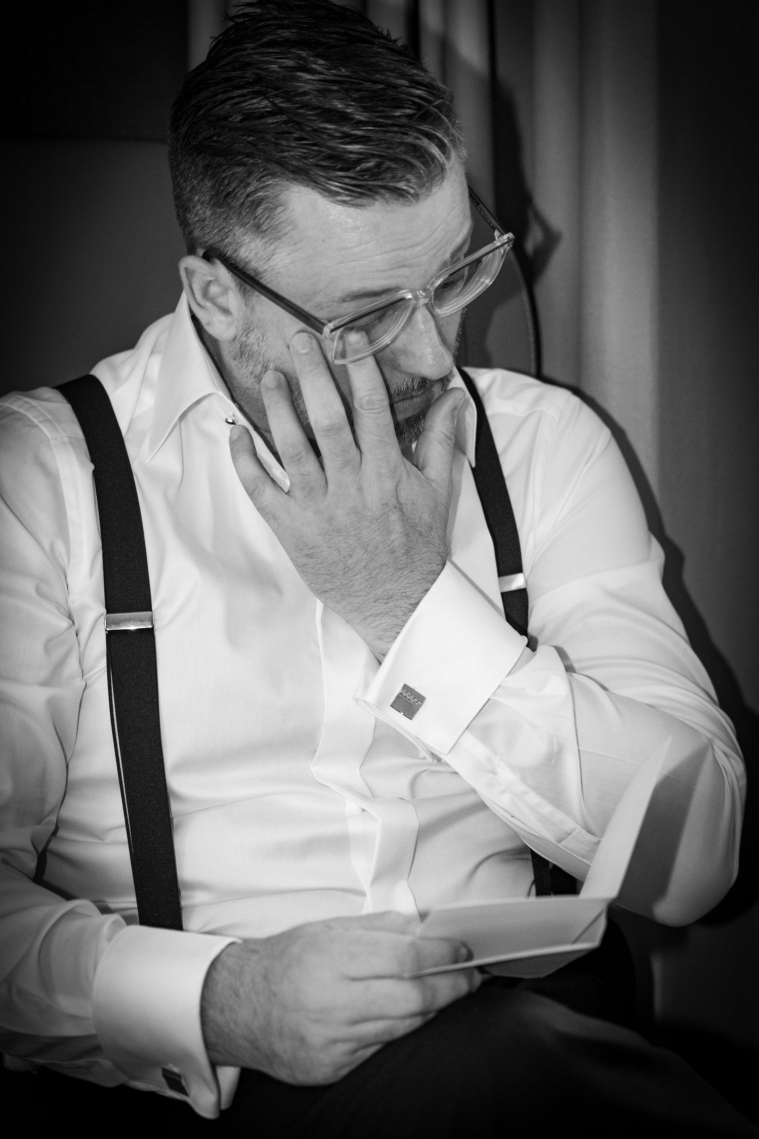 Emotional groom in suspenders reading heartfelt note in black and white wedding portrait.