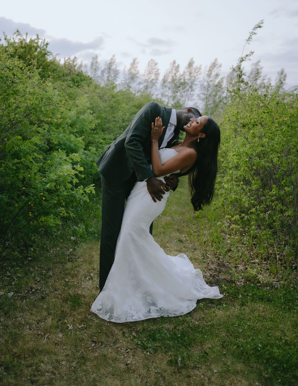 A couple in formal wedding attire share a romantic dip pose together outdoors surrounded by green foliage.