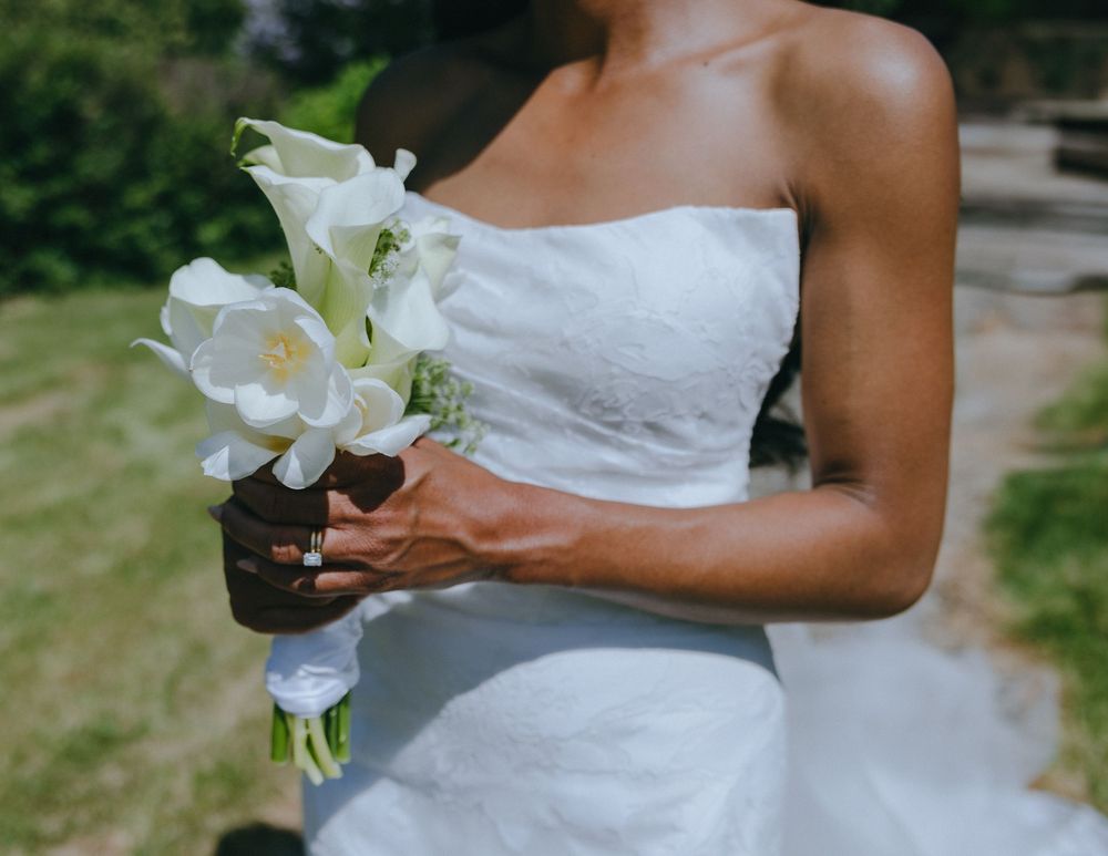 A person in a white strapless wedding dress holds an elegant bouquet of white lilies outdoors in natural light.