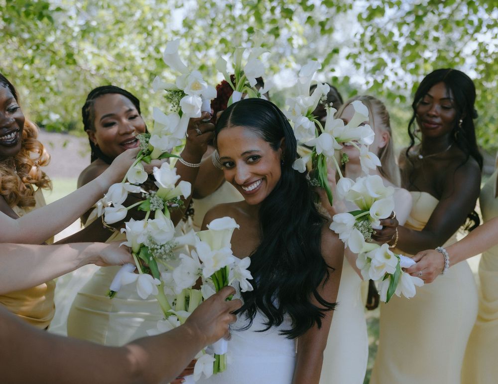A bride in a white dress surrounded by bridesmaids in yellow dresses holding white lily flowers at an outdoor wedding.