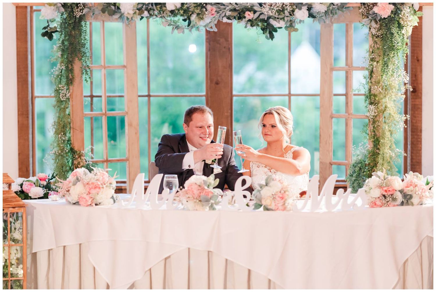 Newlyweds share a candid laughing moment at their wedding reception head table decorated with pink flowers and greenery.