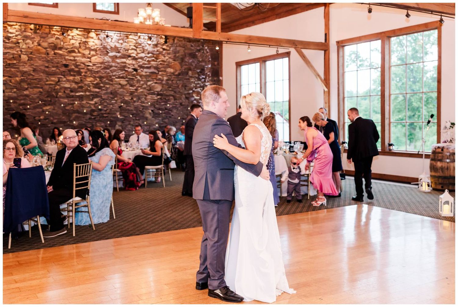 Couple sharing a romantic first dance in a rustic venue with stone walls and natural lighting through large windows.