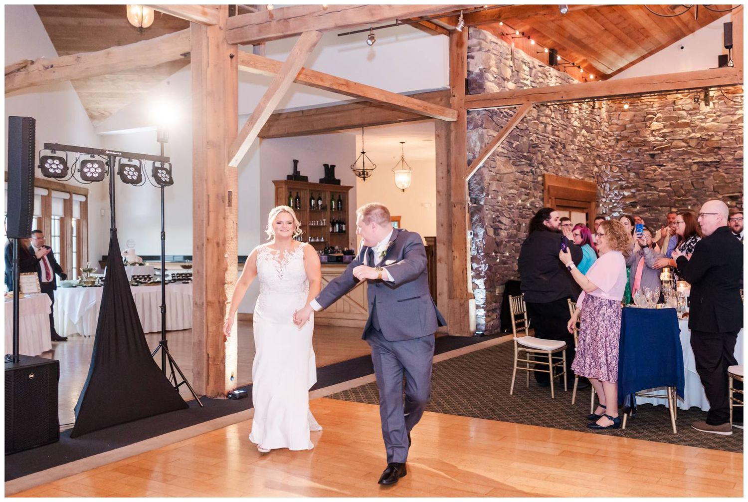 A couple makes their grand entrance at a rustic barn wedding venue with exposed wood beams and stone walls while guests watch.