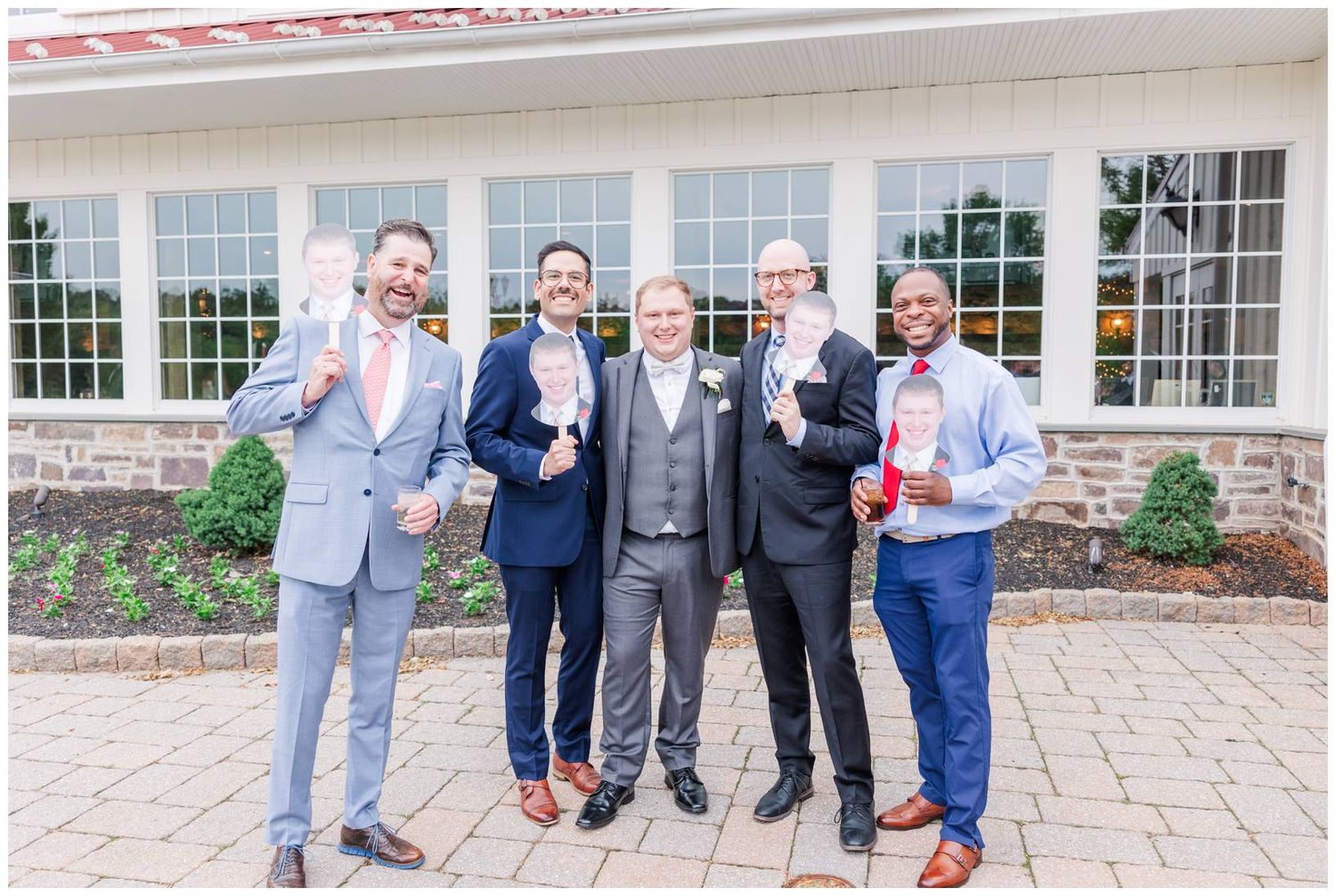 A group wearing suits stands together while holding drinks on a brick patio outside a window-lined building.