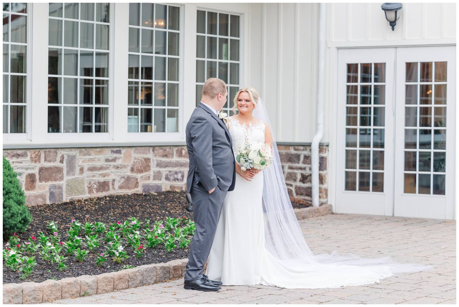 A couple in wedding attire share a moment together outside a stone and white building with french doors and windows.