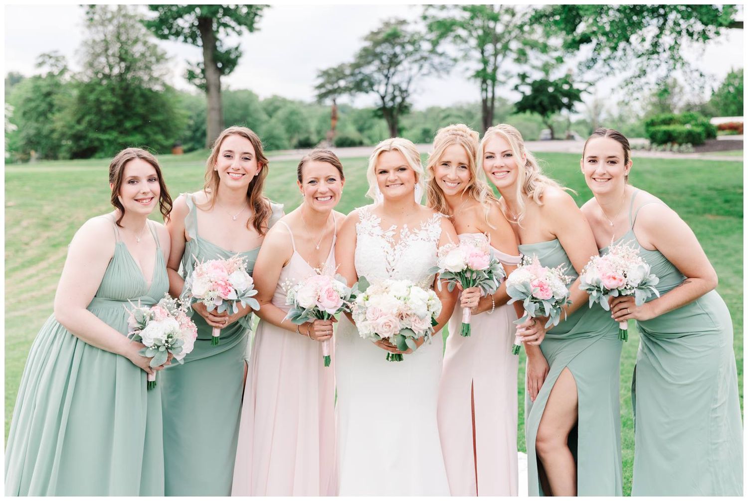 Bridesmaids in soft pink and sage green dresses pose with bouquets on a sunny day.