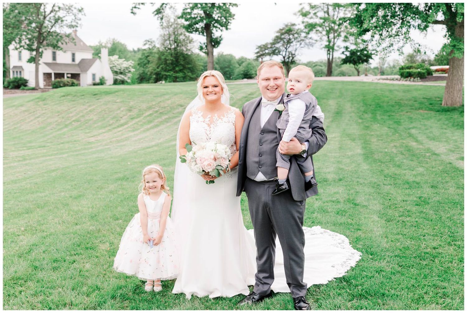 Joyful wedding portrait on a lush green lawn with elegant white gown and gray suit attire.