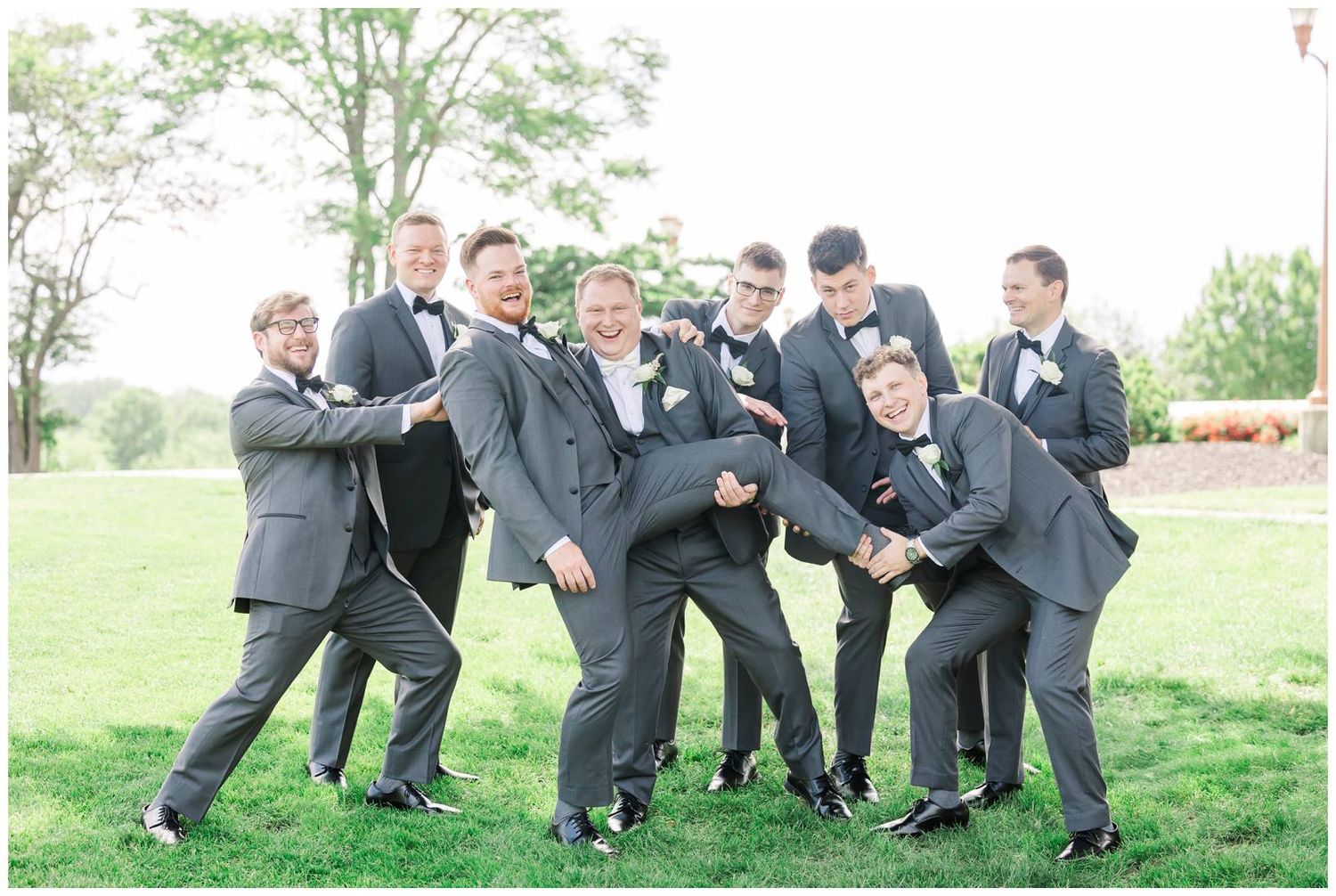Wedding party in matching gray suits playfully pose together on a grassy lawn during outdoor celebration.