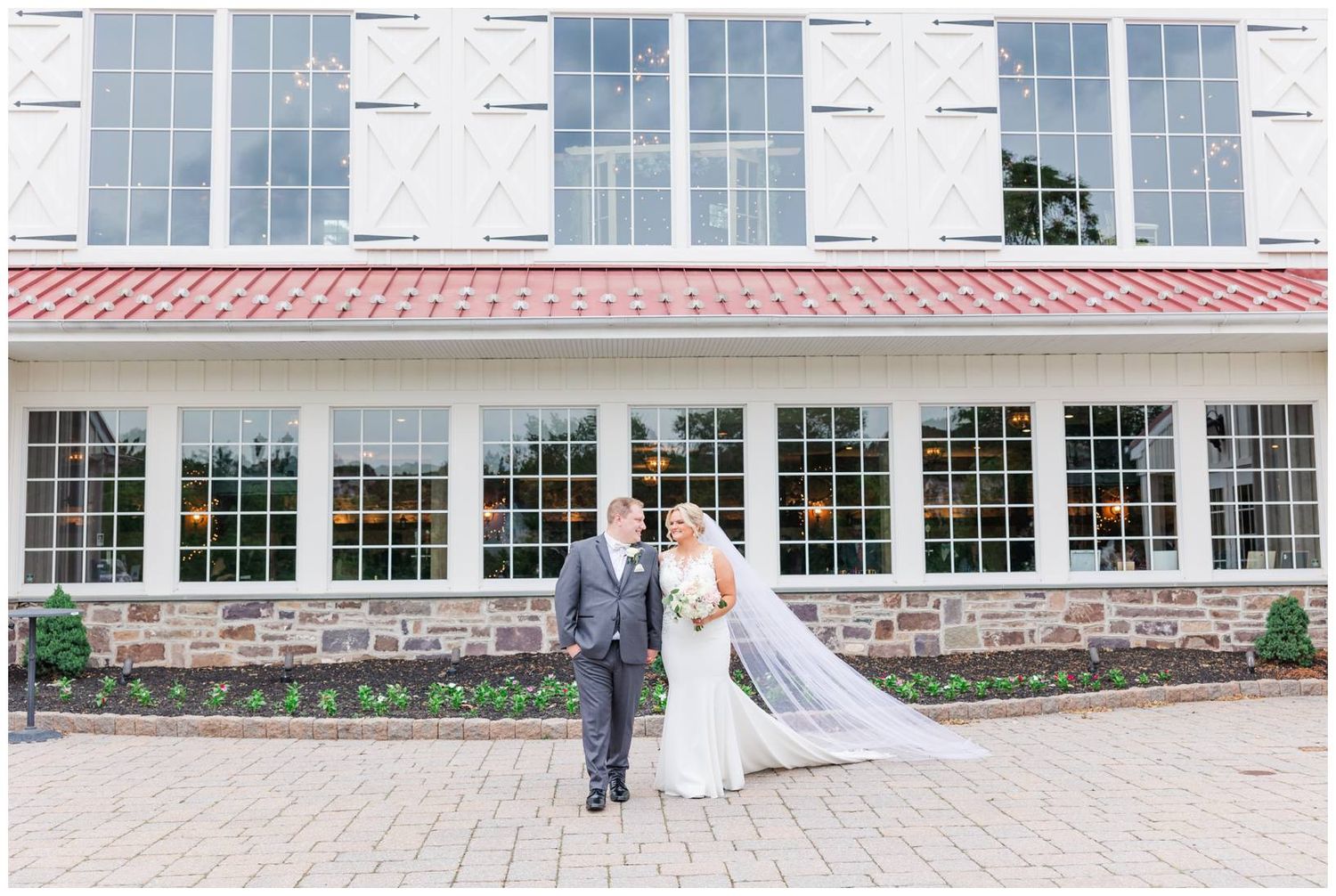 A couple stands in formal attire in front of a white stone building with large windows and a red tiled roof trim.