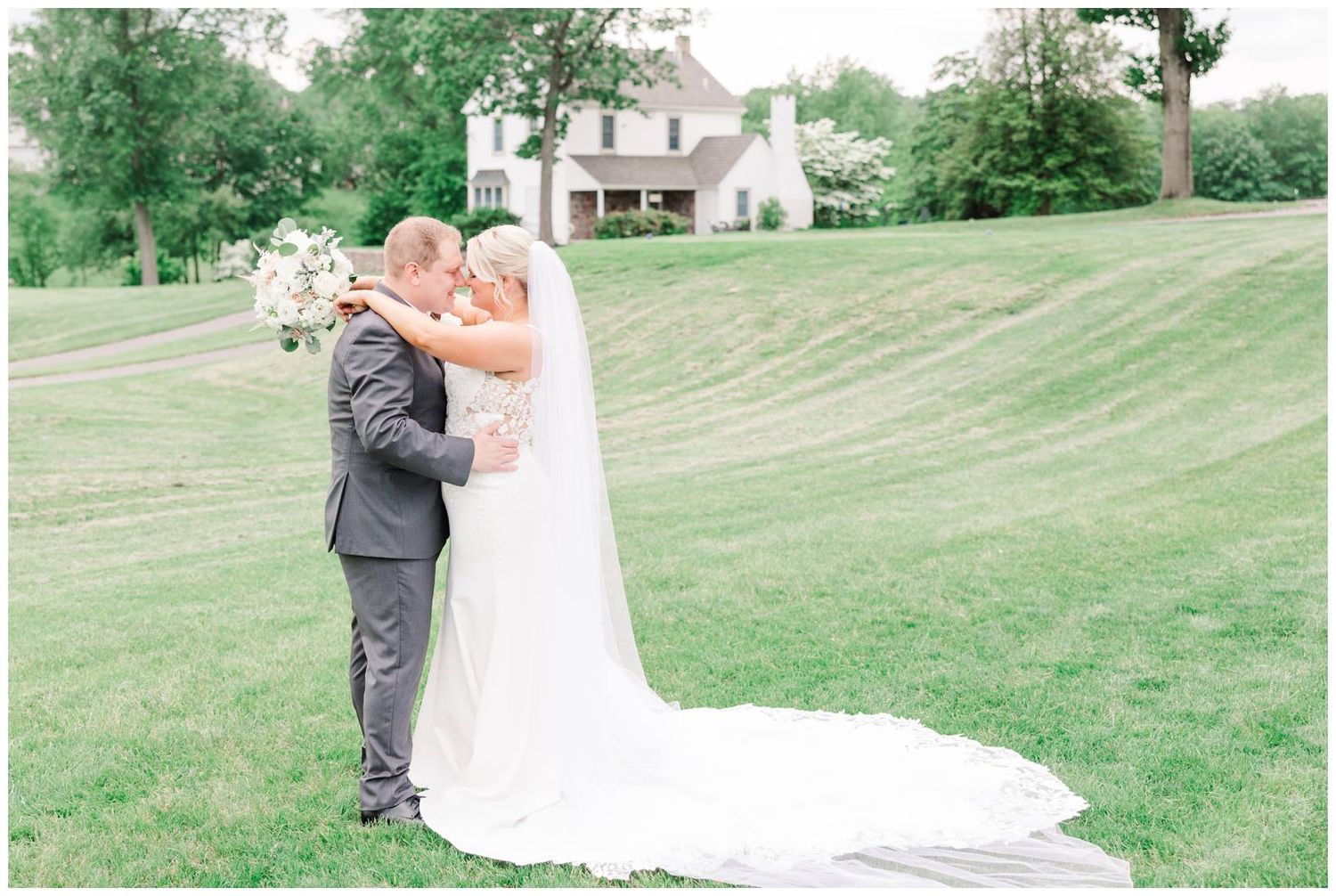 A couple embraces outdoors on their wedding day at a beautiful estate with lush green lawn and white house in background.