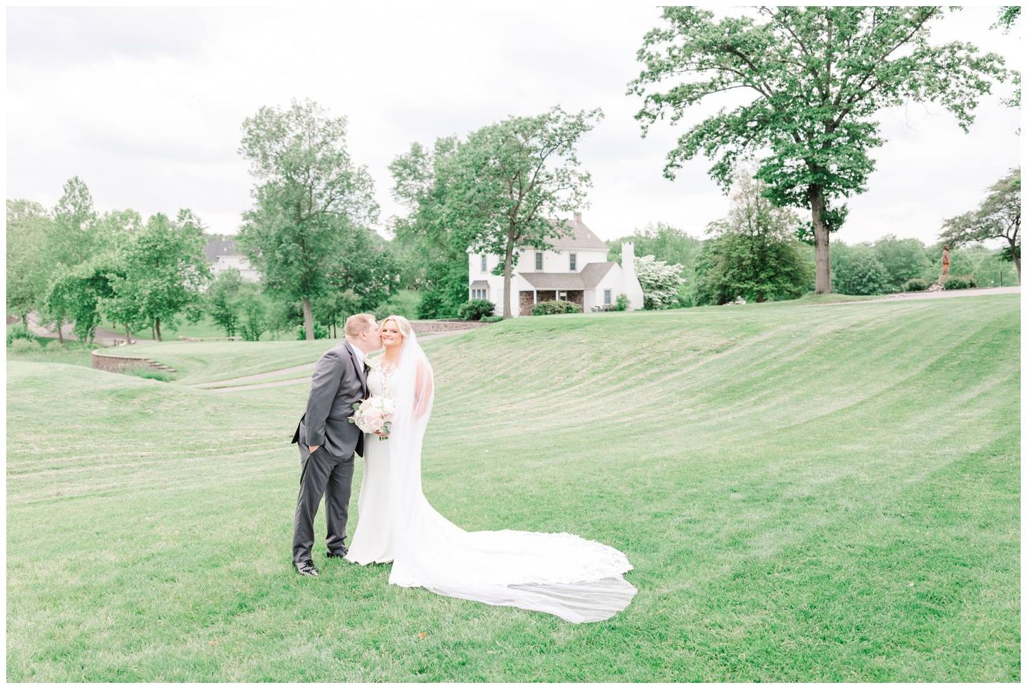 Newlyweds in formal attire stand together on a lush green lawn with a house visible in the background.