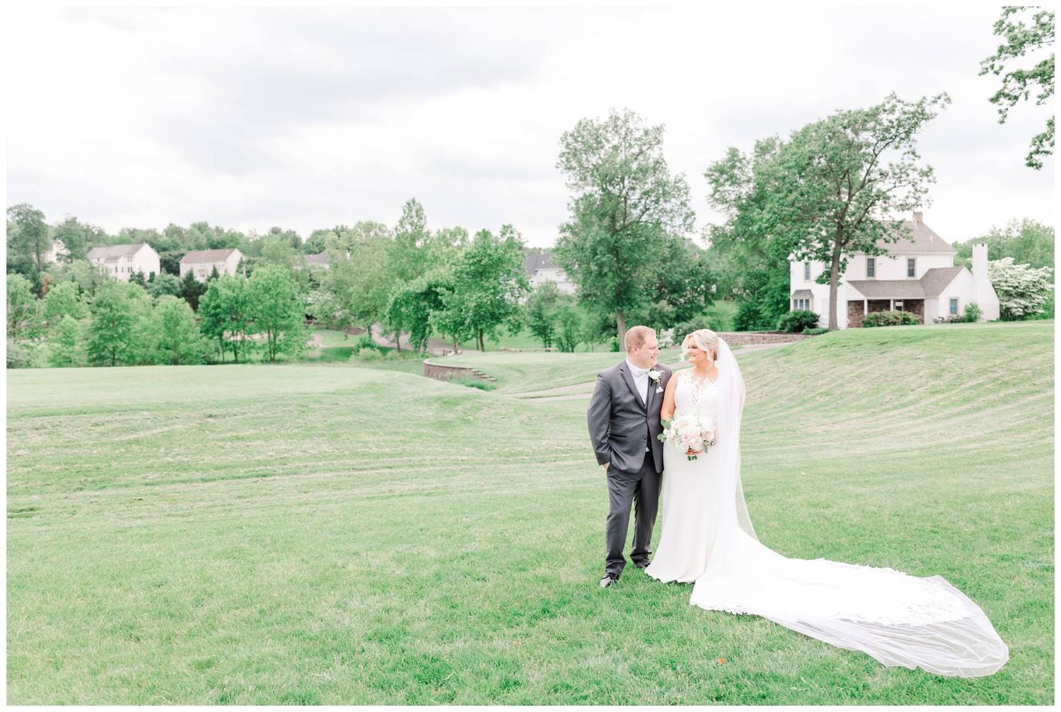 A wedding couple stands on a lush green hillside with white houses and trees in the background.