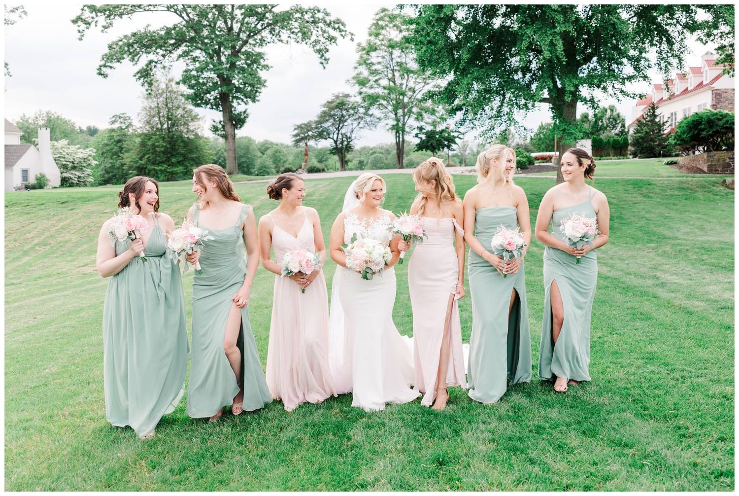 Wedding party in flowing pastel dresses poses together on a lush green lawn during an outdoor celebration.