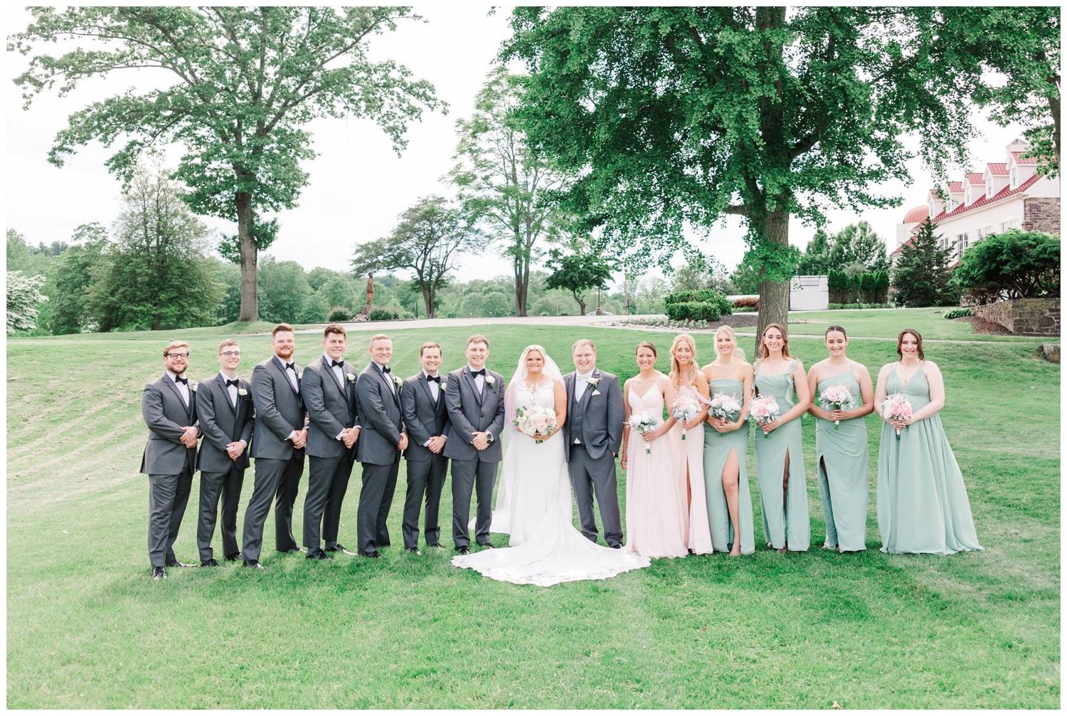 Wedding party in gray suits and pastel dresses pose together on green lawn with trees in background.