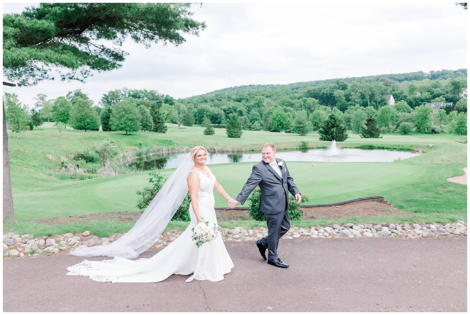 A couple walks hand in hand along a scenic path on a golf course with rolling green hills and trees in the background.