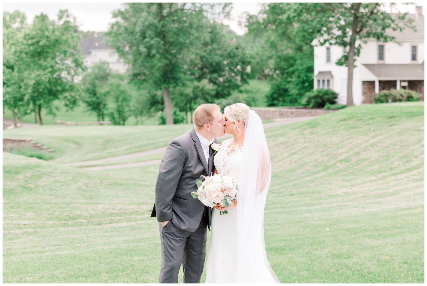 A bride and groom share an intimate moment on a lush green hillside with white buildings in the background.