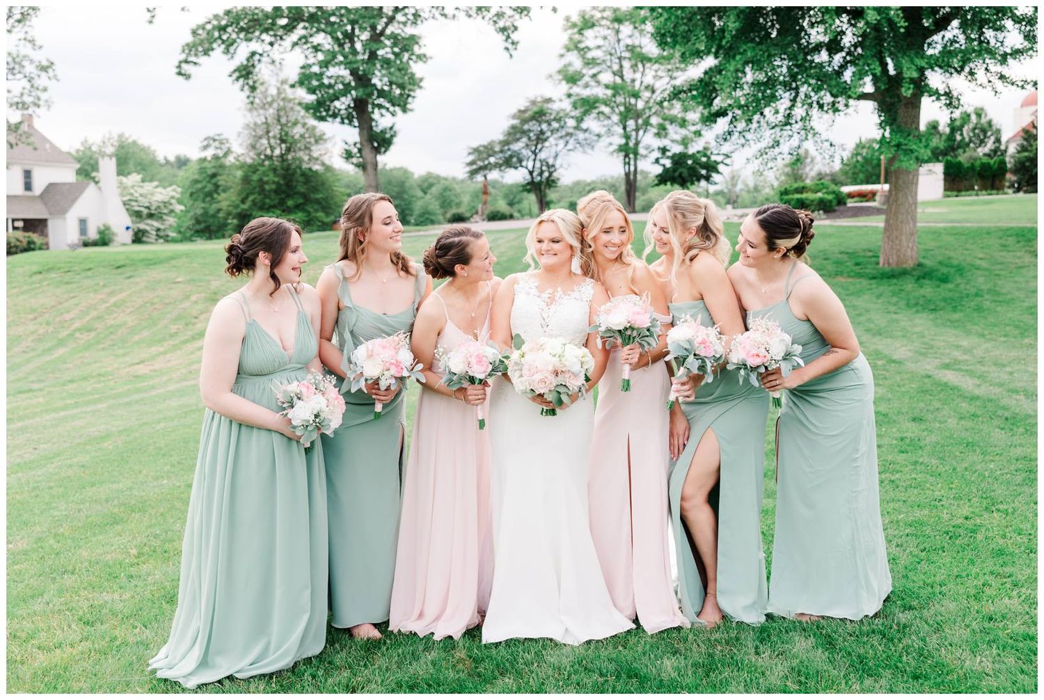 Group in pastel green and pink bridesmaid dresses holding bouquets while smiling together on a grassy lawn.