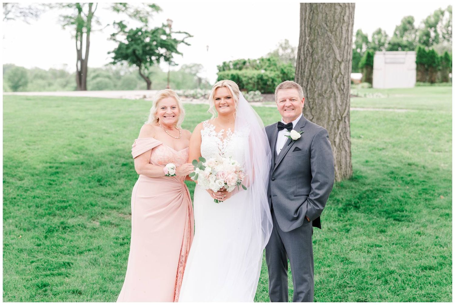 Wedding party poses together on a sunny day in a grassy outdoor setting under a large tree.