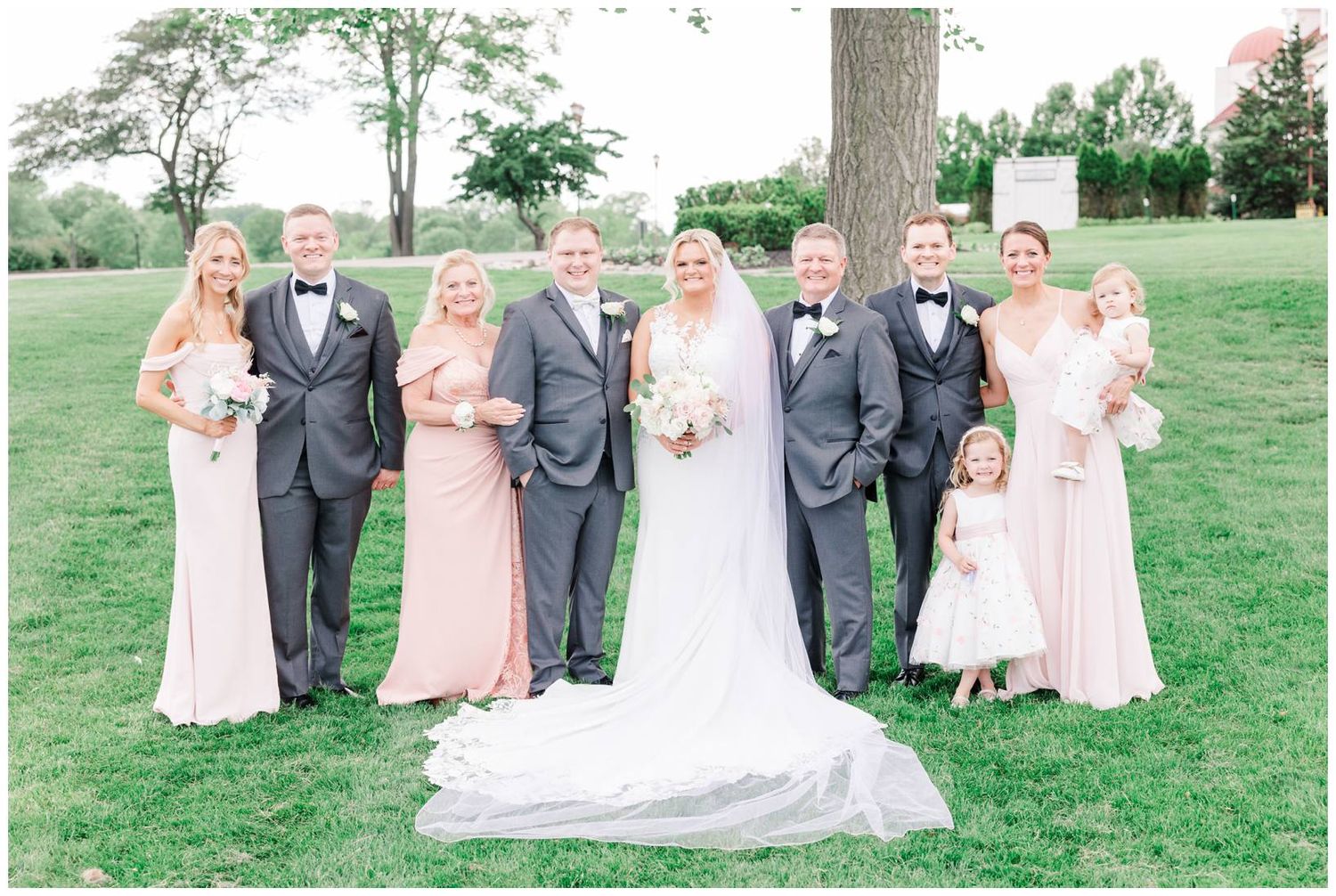 A wedding party stands outdoors on a lush green lawn wearing blush pink and gray formalwear on a spring day.