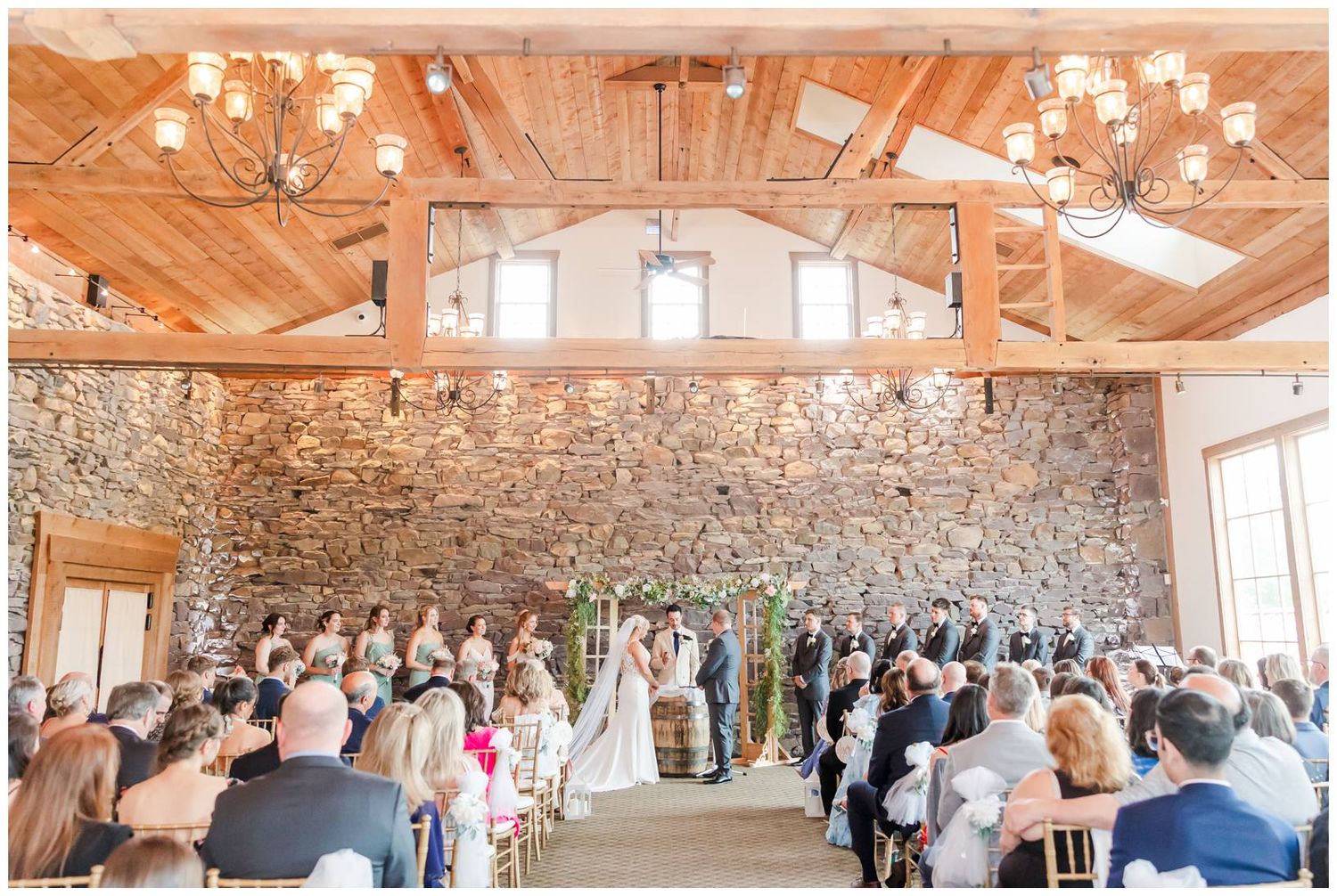 Wedding ceremony in rustic venue with exposed stone wall, wooden beams and chandeliers as guests witness special moment.