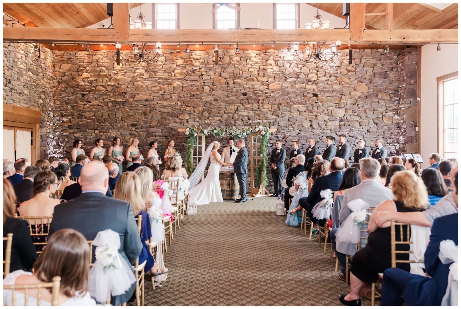 Indoor wedding ceremony at rustic venue with exposed stone walls, wooden beams, and rows of guests seated on chairs.