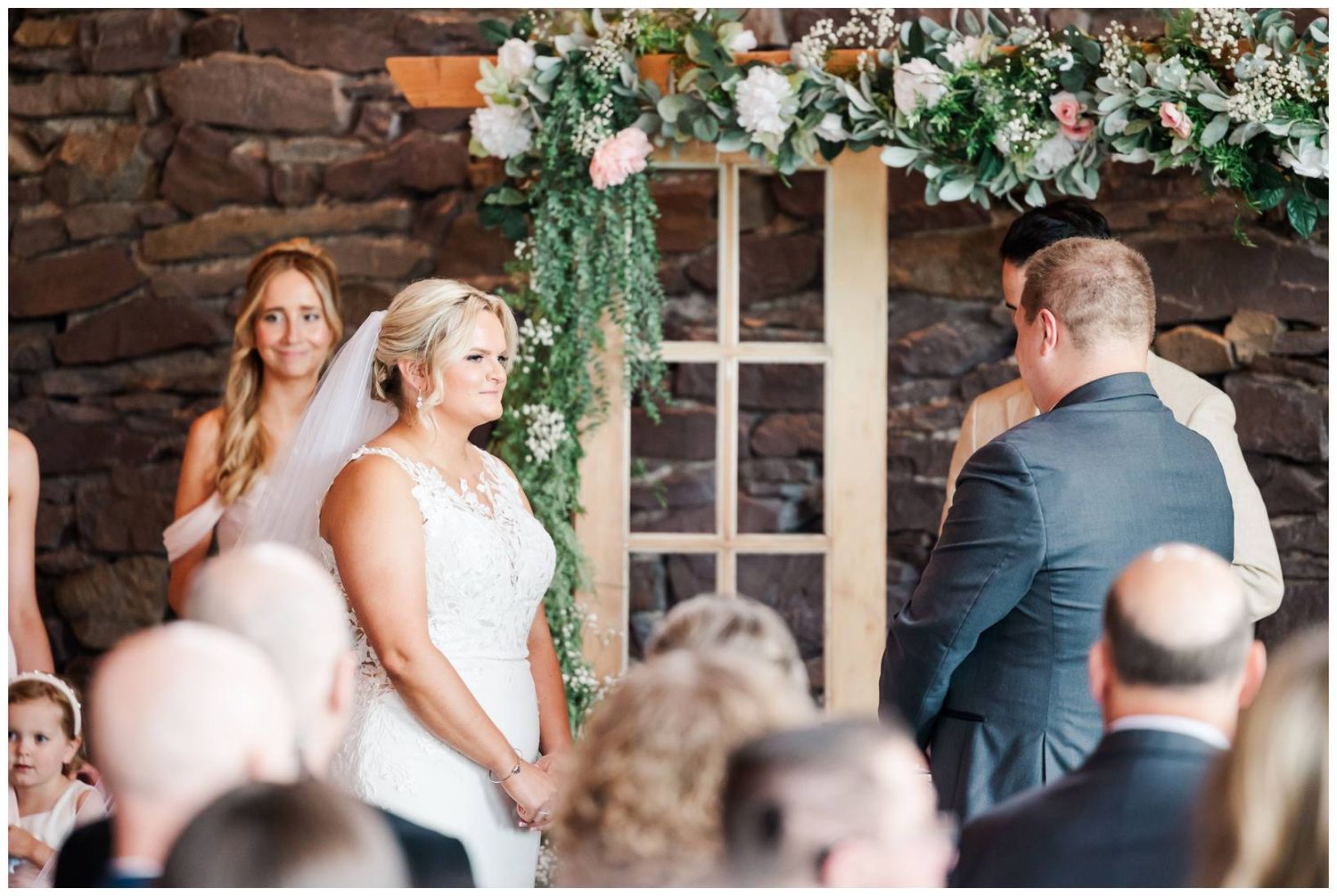 A joyous moment during a wedding ceremony under a floral arch against a rustic brick wall.