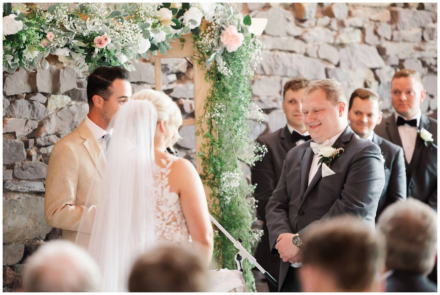 A romantic outdoor wedding ceremony under a floral arch with white blooms against a rustic stone wall.