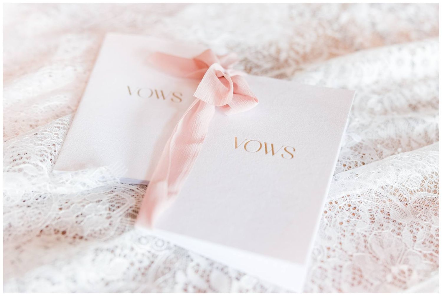 Two white vow books with gold lettering tied with blush pink ribbon rest on delicate lace fabric.