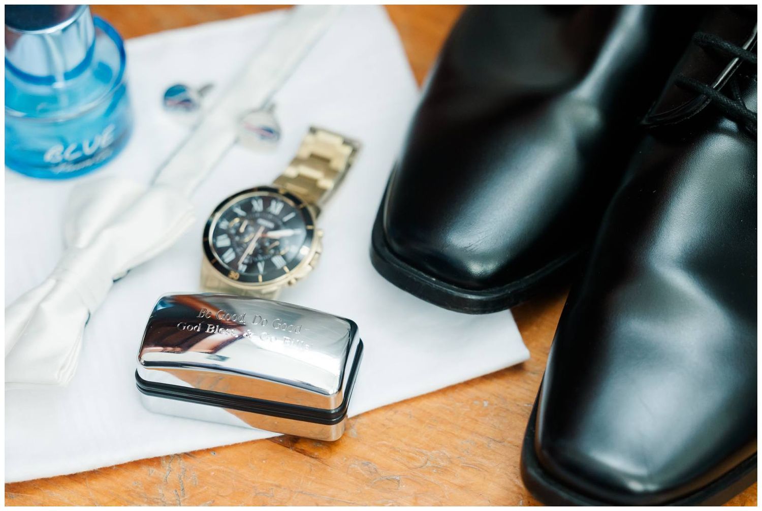 Sleek black dress shoes alongside a silver lighter, wristwatch, and white fabric on a wooden surface.