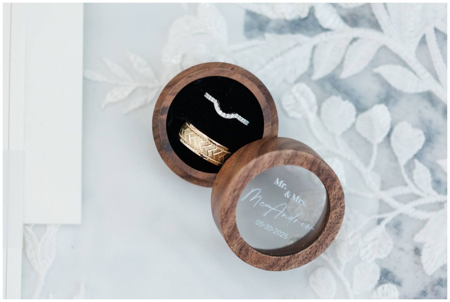 A circular wooden ring box with a dark interior is displayed on white floral paper with elegant styling.