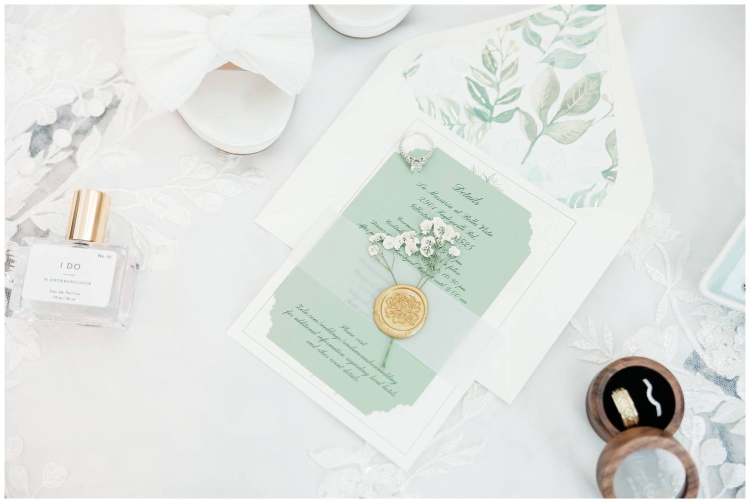 Mint green wedding invitation with botanical details lies on marble surface alongside perfume bottle and decorative elements.
