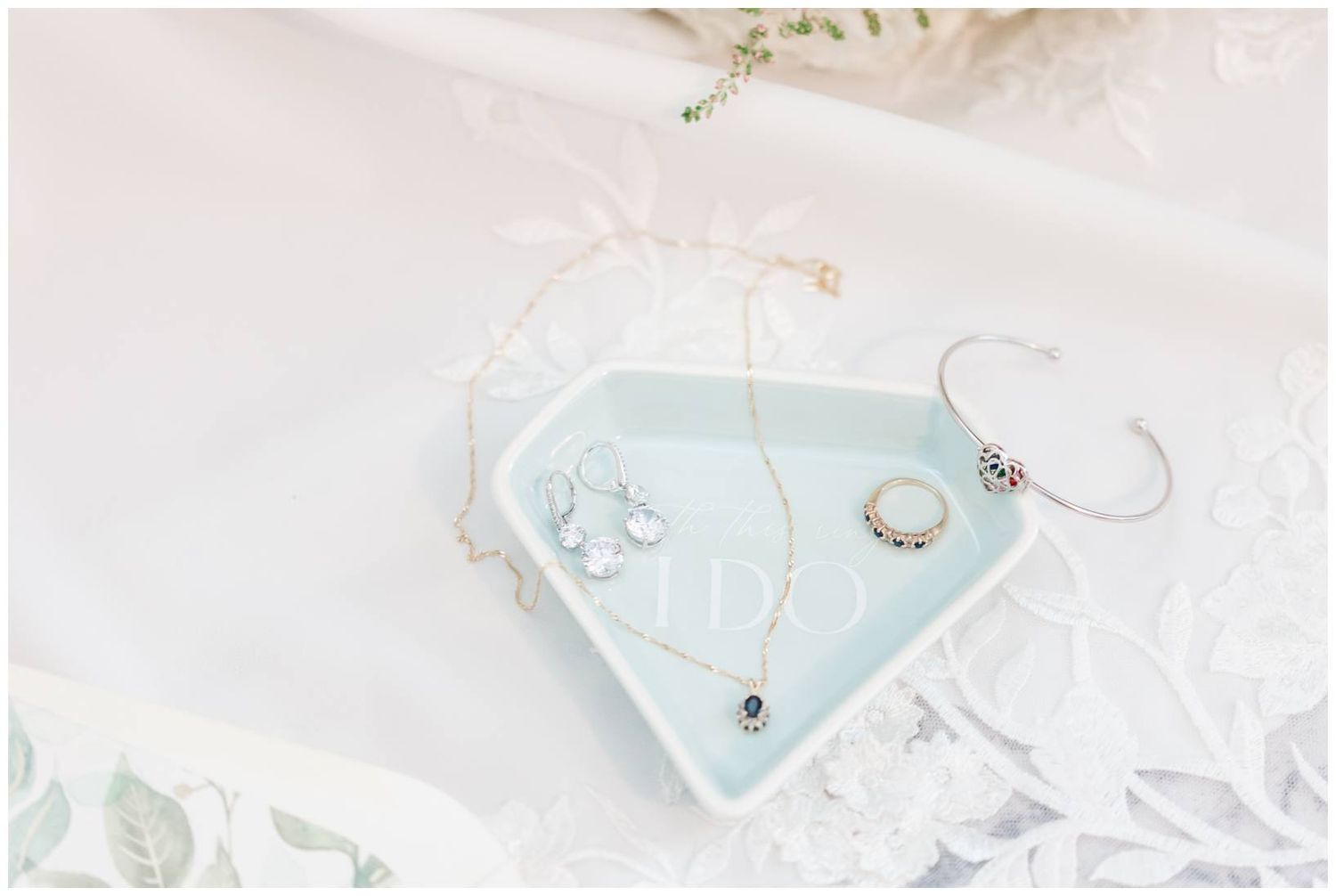 A light blue ceramic diamond-shaped jewelry dish holds delicate rings and earrings against a white lace background.