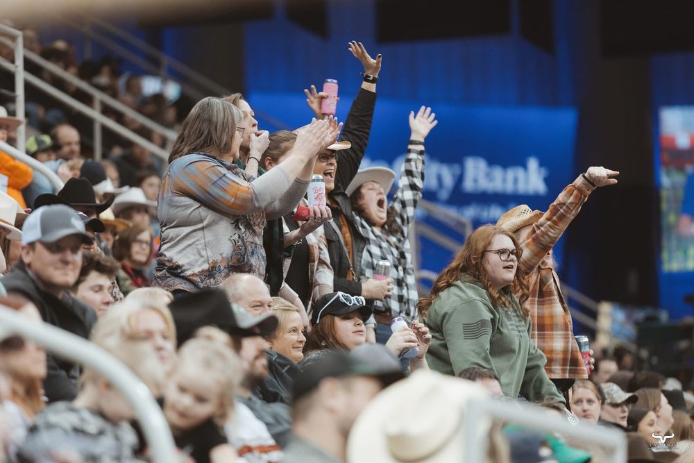 Enthusiastic crowd cheering at rodeo event with raised hands in stadium seating.